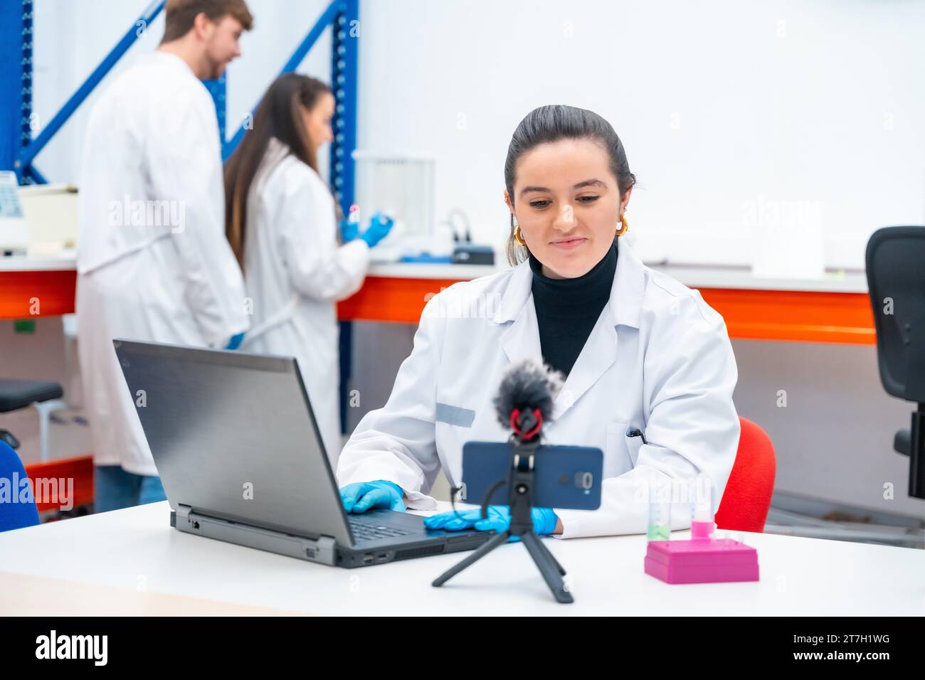 Young female scientist recording video with the mobile while using laptop in a research laboratory Stock Photo