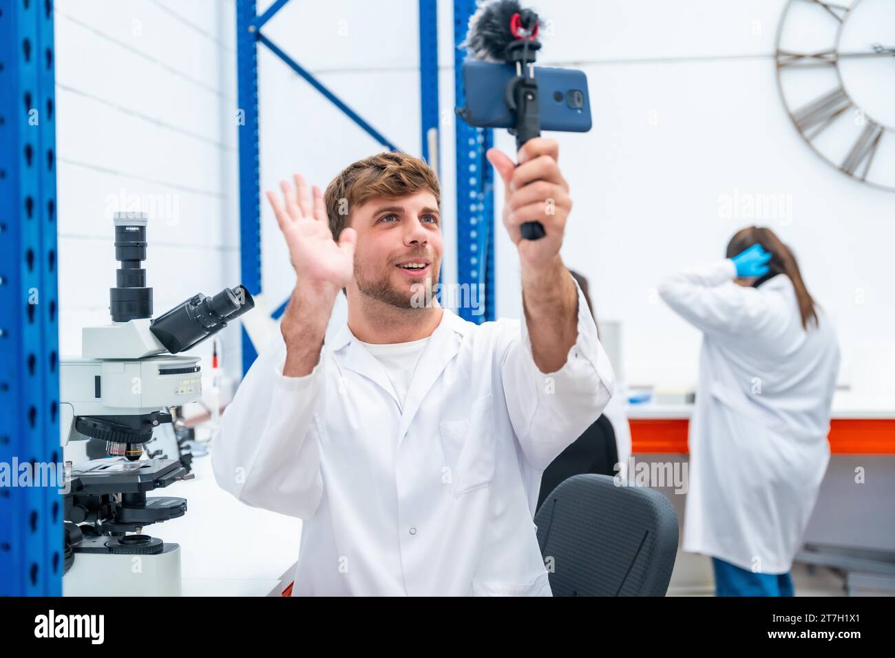 Young handsome scientist waving while broadcasting using phone in a research laboratory Stock Photo