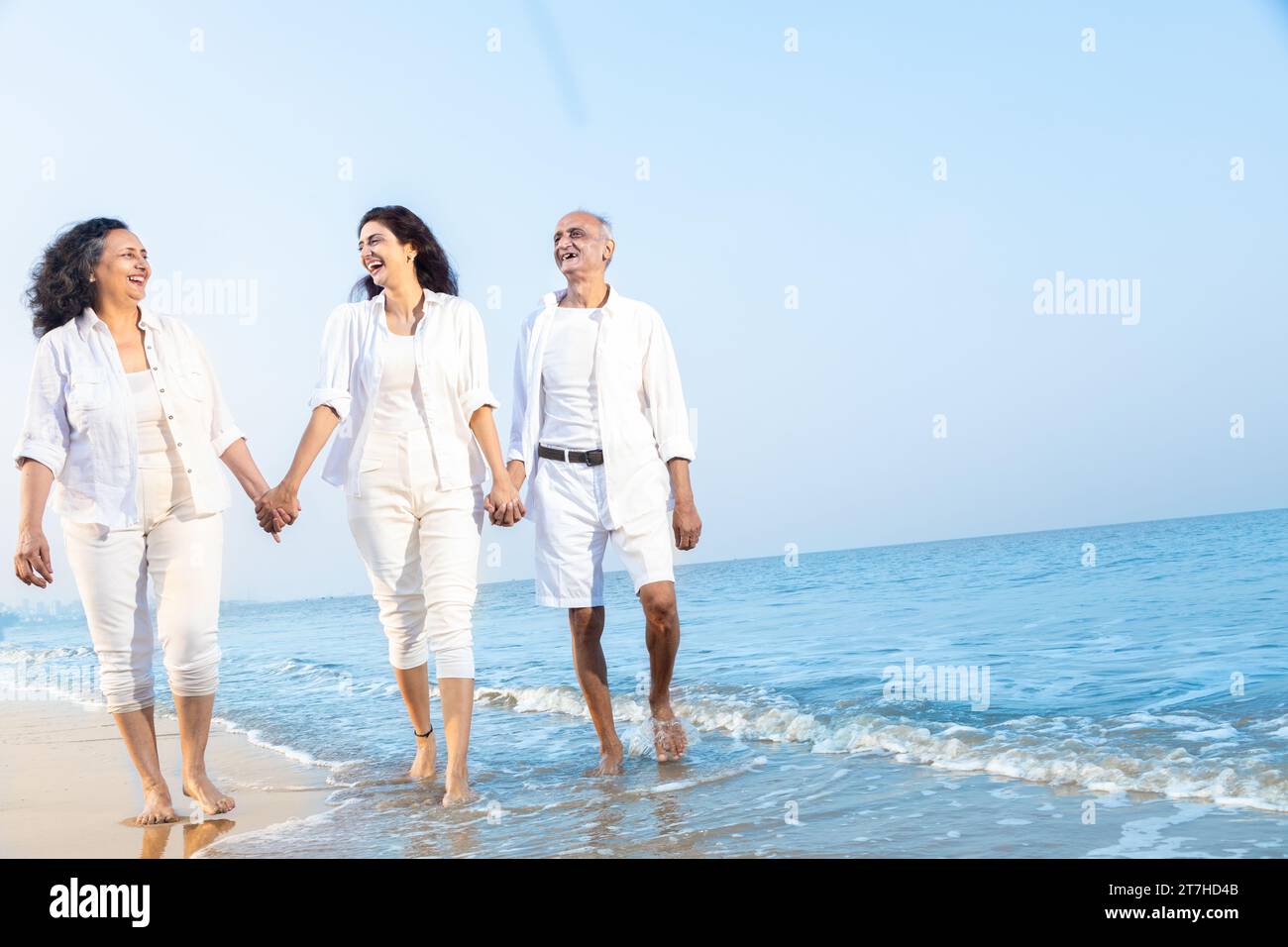 Happy senior indian couple with beautiful young daughter walking and enjoying vacation at beach. Family wearing white cloths having fun together. Stock Photo