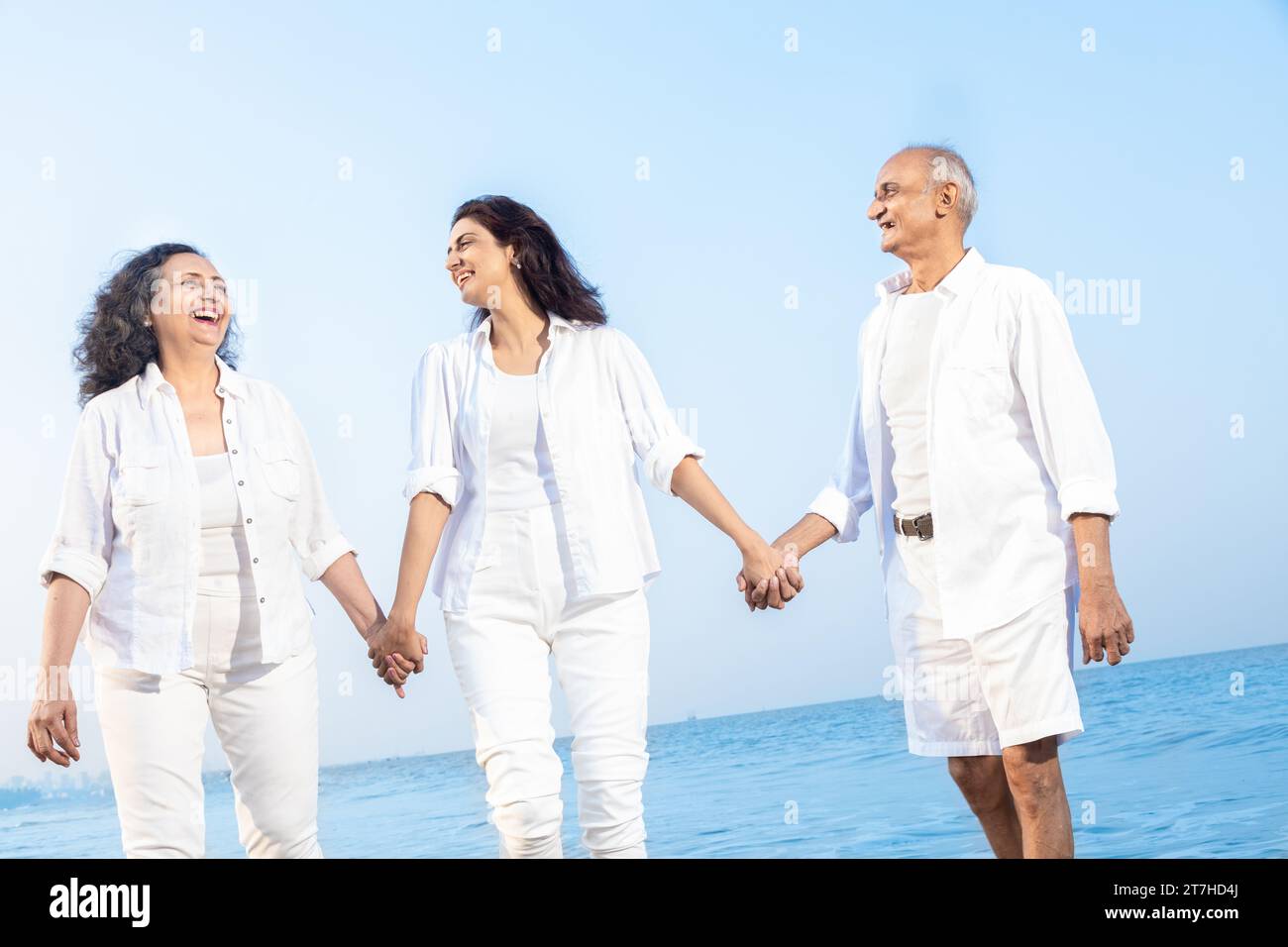 Happy senior indian couple with beautiful young daughter walking and enjoying vacation at beach. Family wearing white cloths having fun together. Stock Photo