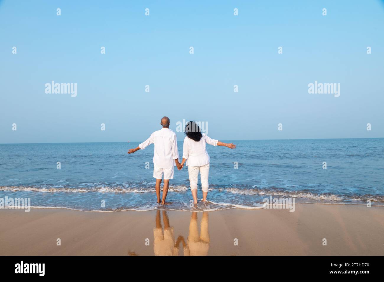 Happy senior indian couple wearing white cloths enjoying summer vacation, holiday at beach looking at sea with open arms Copy space Stock Photo