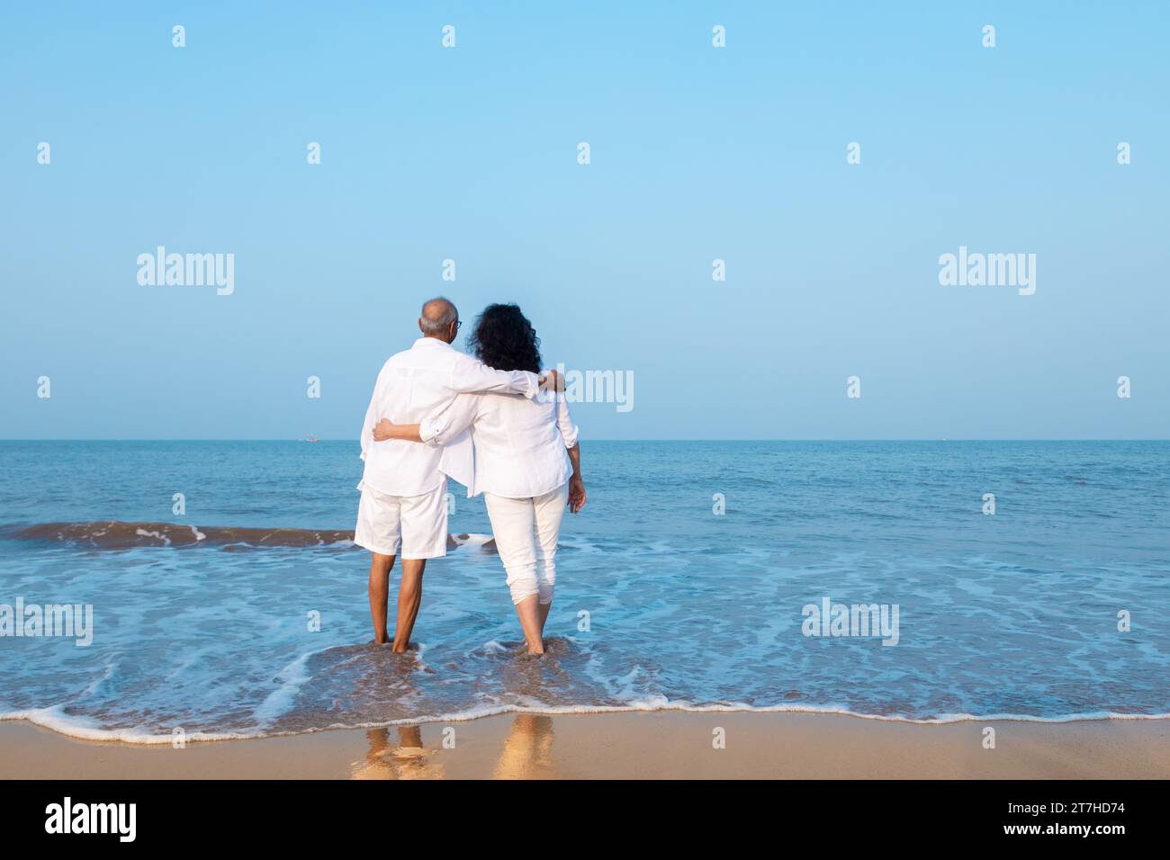 Happy senior indian couple wearing white cloths enjoying summer vacation, holiday at beach looking at sea. Copy space Stock Photo