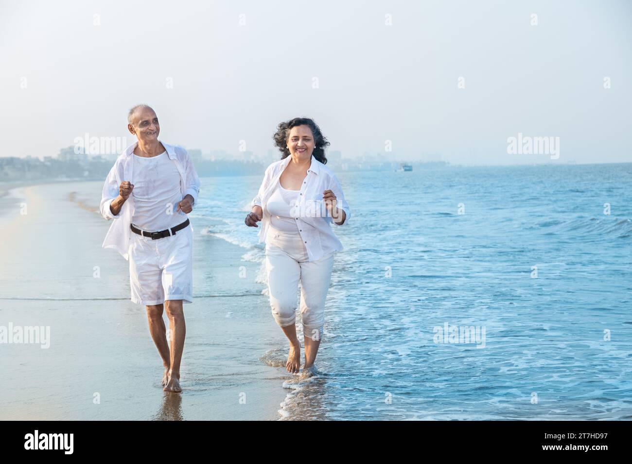 Happy senior indian couple wearing white cloths running together at beach and having fun. Vacations and Health Concept Stock Photo