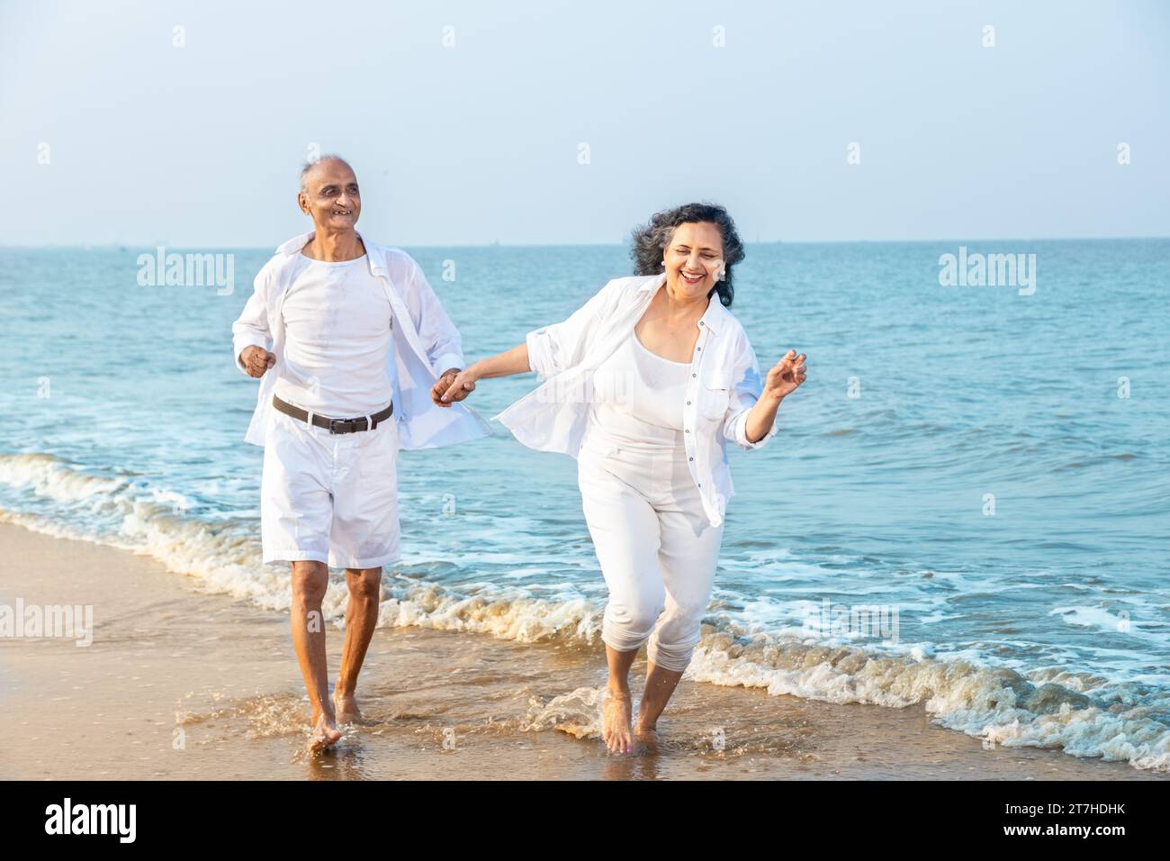 Happy romantic senior indian couple wearing white cloths running together and having fun at beach , carefree life, Vacations and health concept Stock Photo