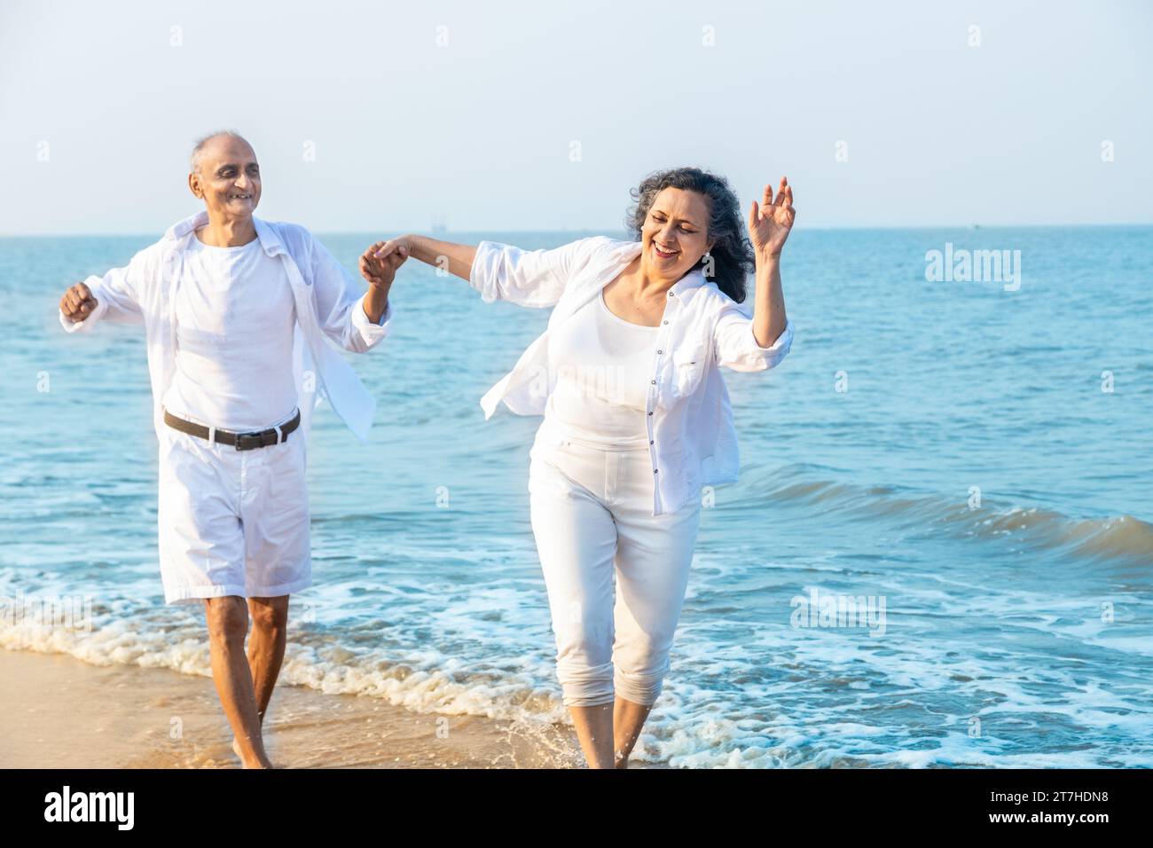 Happy romantic senior indian couple wearing white cloths running together and having fun at beach , carefree life, Vacations and health concept Stock Photo