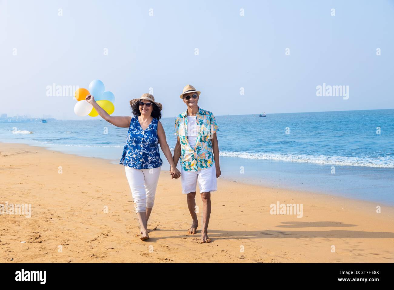 Senior indian couple with colorful balloon in hands walking together at the beach. Enjoying vacation, holiday at beach. Copy space. Stock Photo