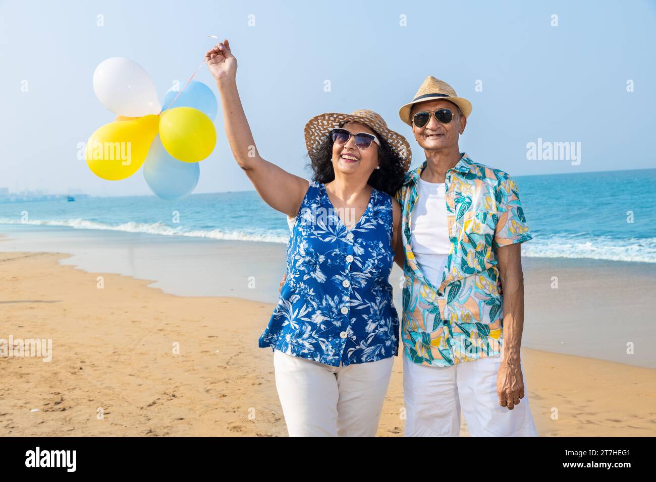 Senior indian couple with colorful balloon in hands walking together at the beach. Enjoying vacation, holiday at beach. Copy space. Retirement life. Stock Photo