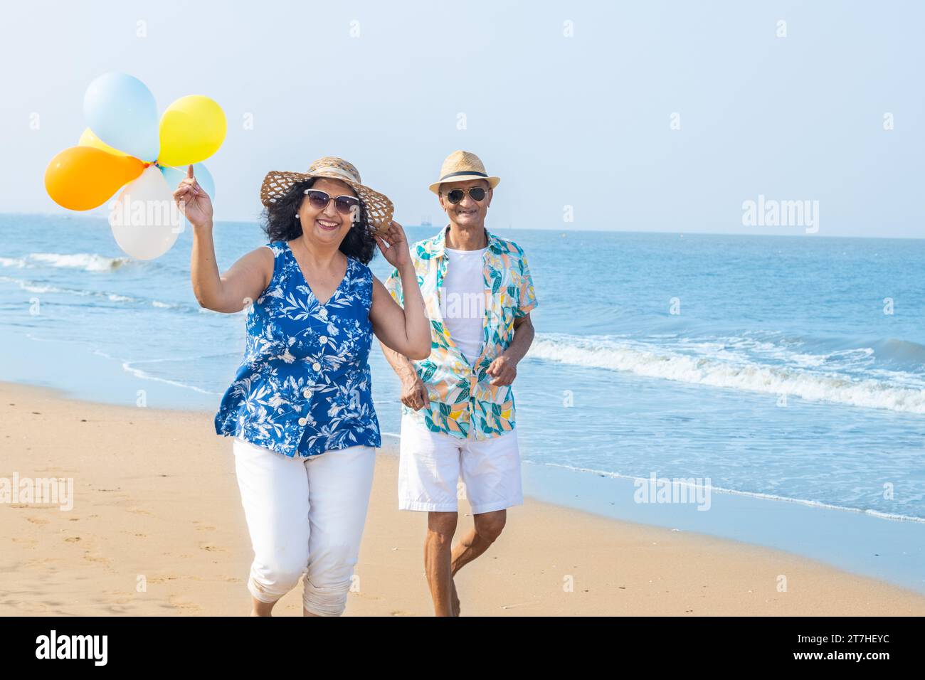 Senior indian couple with colorful balloon in hands running together at the beach. Enjoying vacation, holiday at beach. Copy space. Retirement life. Stock Photo