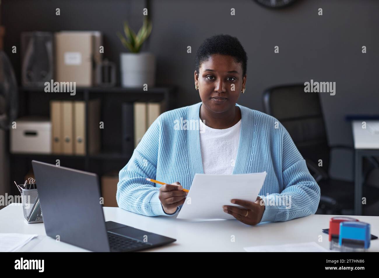 Young confident female manager of visa application center in casualwear looking at camera while sitting by workplace and checking data Stock Photo