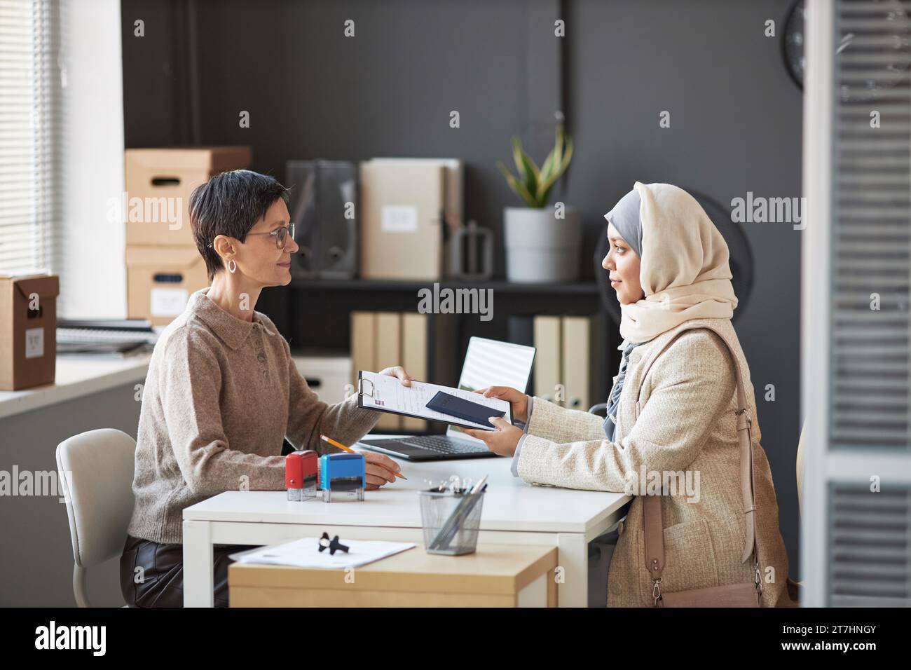 Side view of mature manager of visa application center and young Muslim female applicant wearing hijab and quiet luxury attire Stock Photo