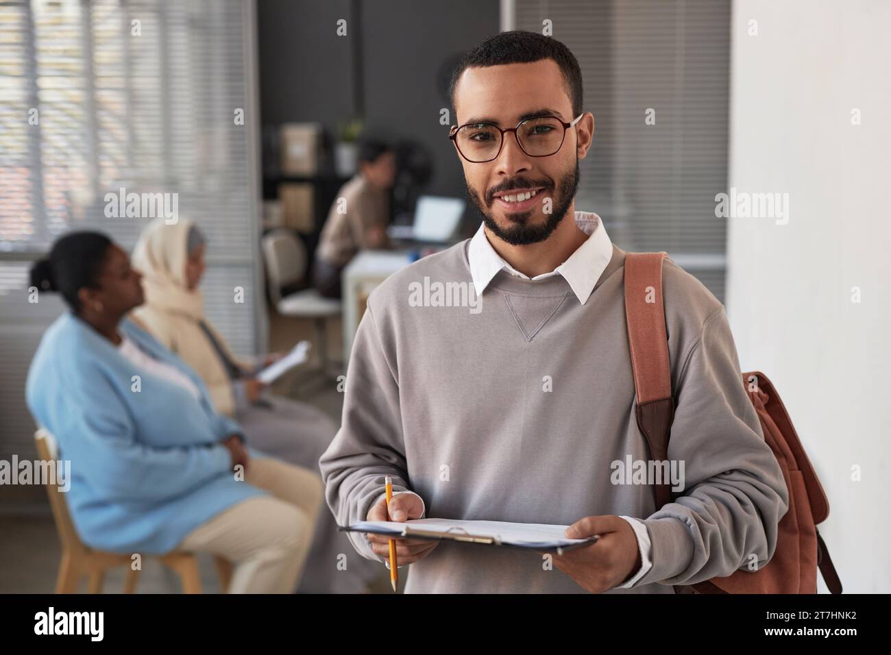 Young smiling multiethnic man with document looking at camera while filling in visa application form and waiting for his turn Stock Photo
