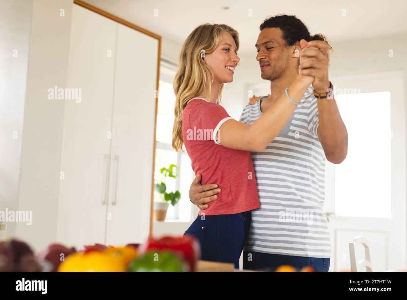 Happy diverse couple dancing at counter with vegetables in sunny