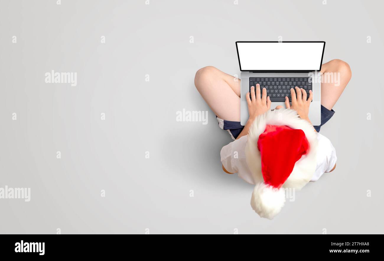 Boy in red Christmas hat works on laptop with isolated screen for mockup. Festive productivity on the floor. Copy space Stock Photo