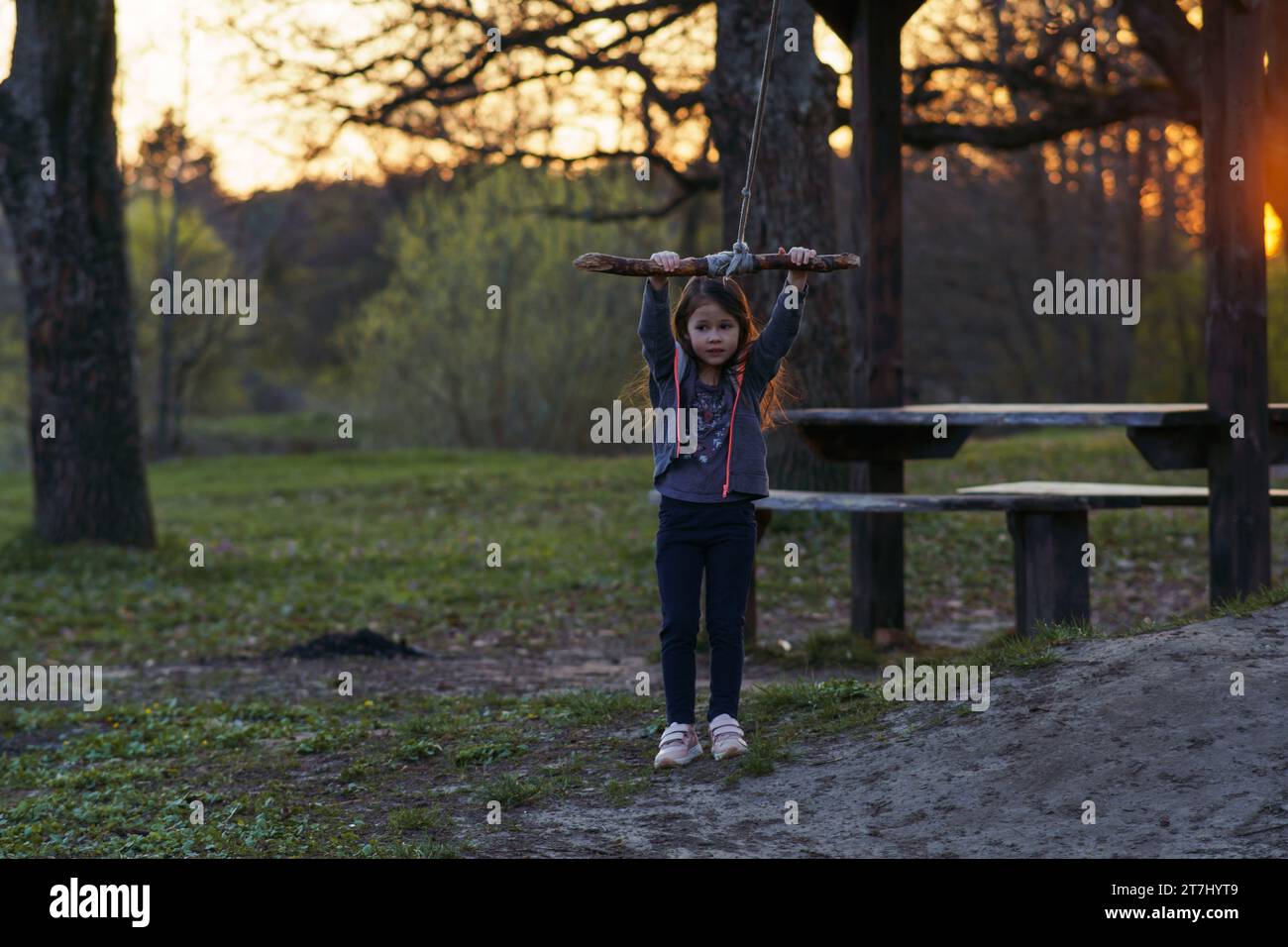 Charming little girl is going to swing on homemade swing in nature, in park, in village, in children's camp. Outdoor games and entertainment. Happy ch Stock Photo