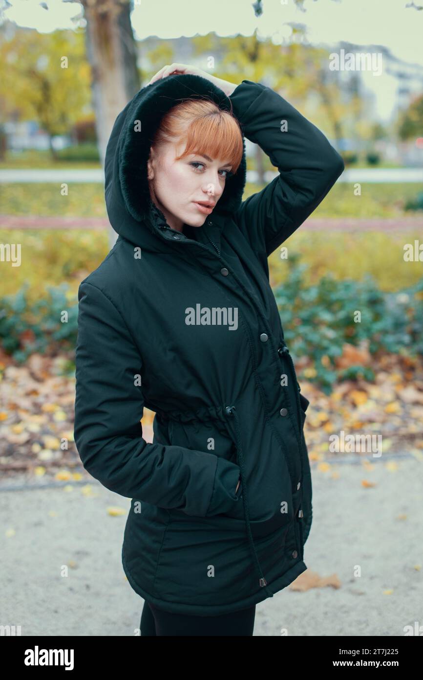 Redhead woman in hooded black coat posing at autumn during forest walk Stock Photo