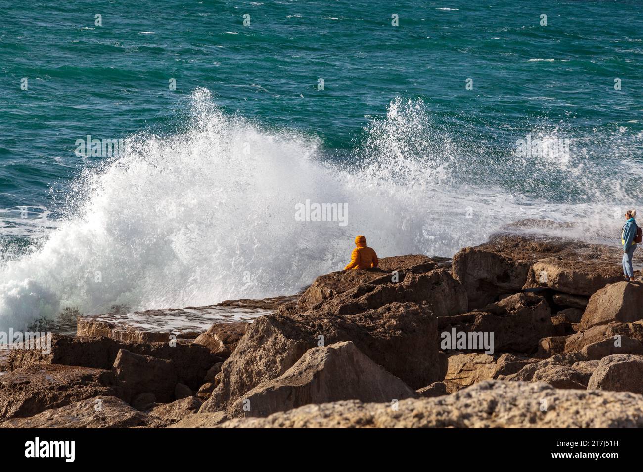 Portland Bill, Isle of Portland, Dorset, England, UK: person sits on Portland stone platform with waves crashing in. second person watches from behind Stock Photo