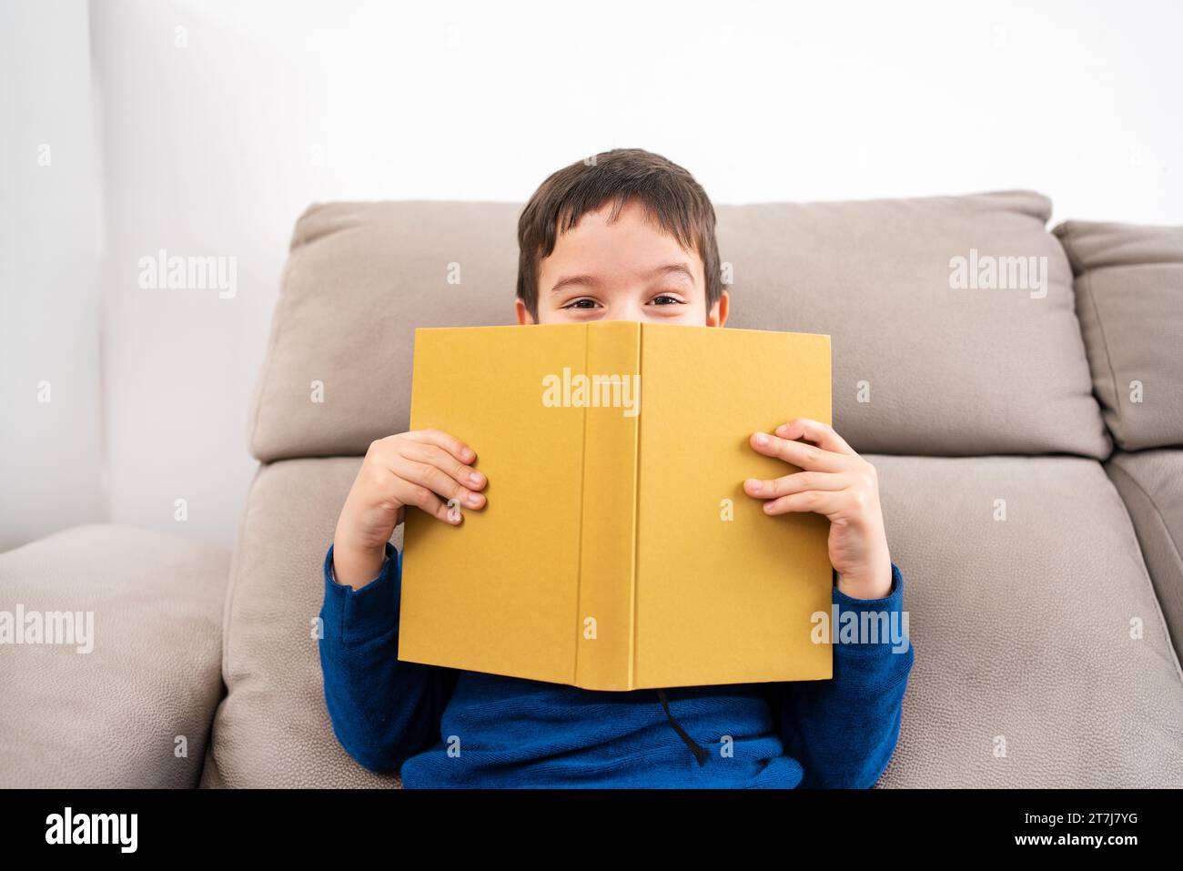 Child sitting on sofa and holding book in front of his face at living room Stock Photo