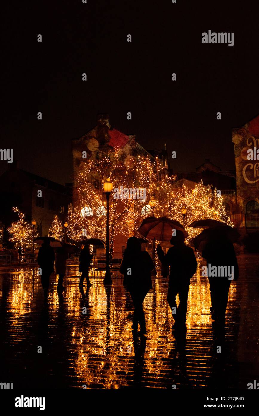 Enchanting Christmas scene: Rain-kissed lights adorn trees, casting reflections on the wet ground as silhouettes of people stroll through the festive Stock Photo