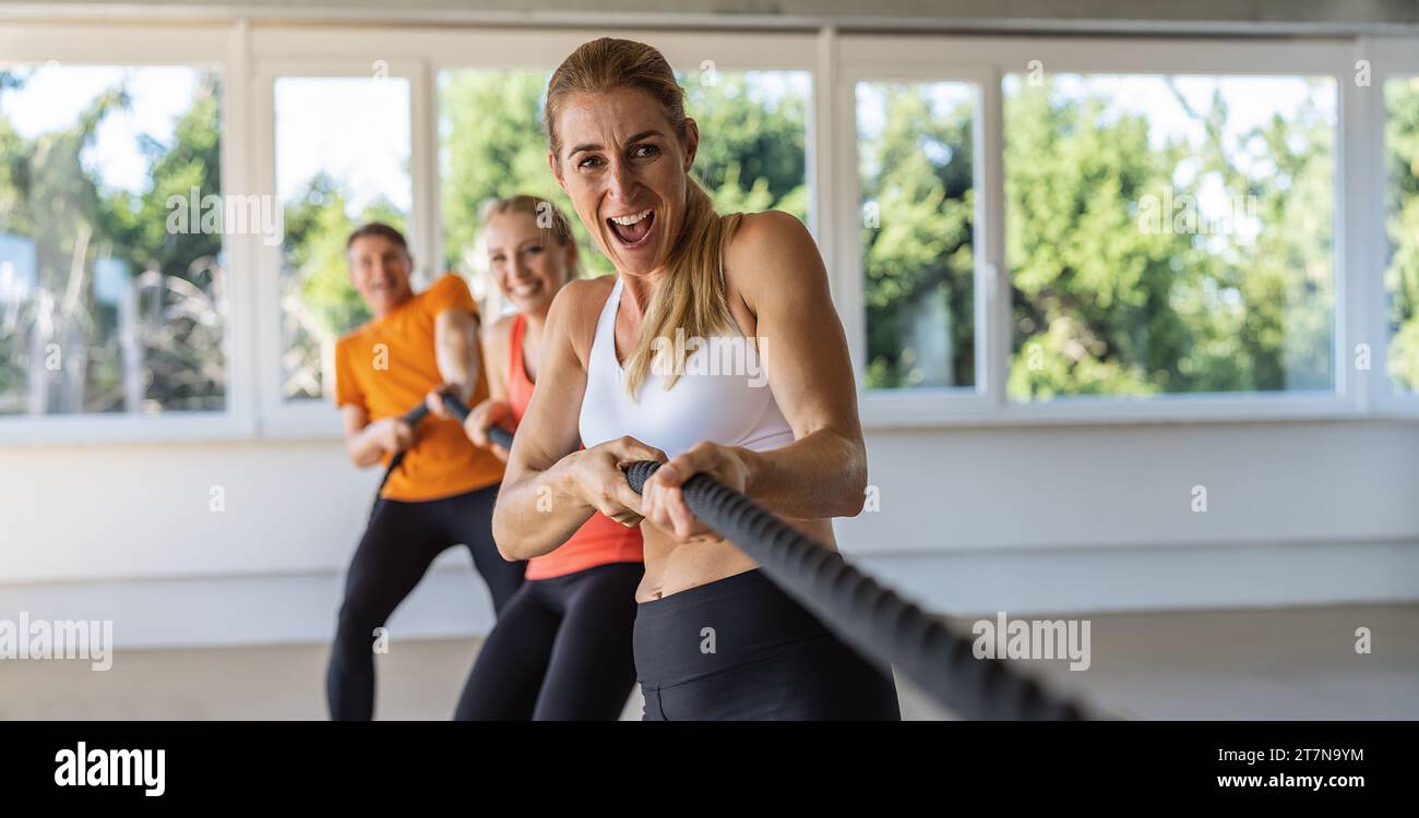 woman pulls on a rope with expressive shout as tug of war competition in a group as teambuilding exercise. Gym for fitness exercises concept image, ba Stock Photo