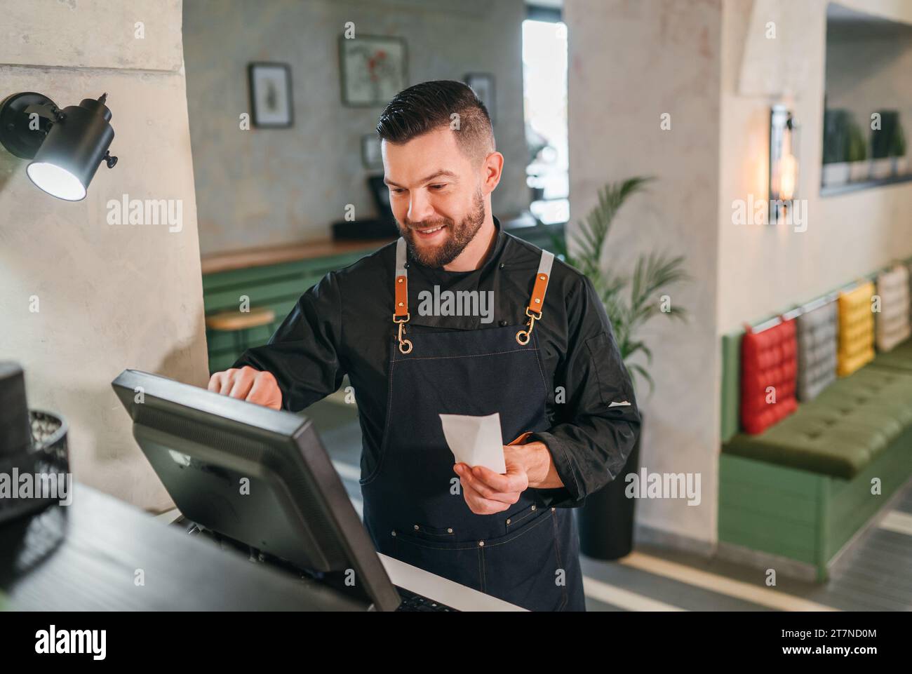 Stylish bearded smiling waiter dressed black uniform processing customer orders using point of sale order terminal system touch screen. Successful peo Stock Photo