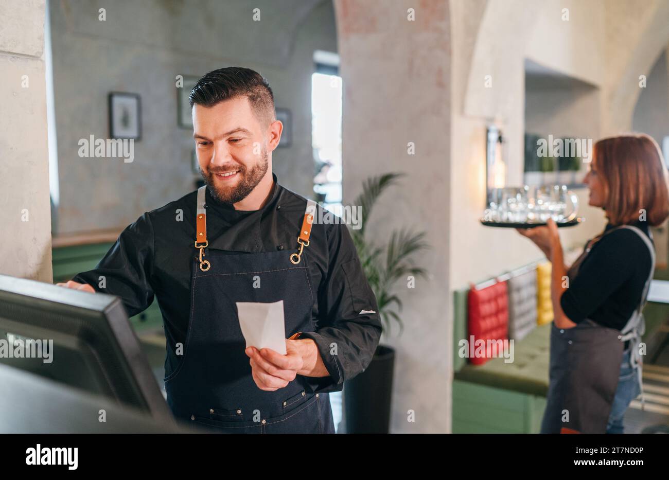 Stylish bearded smiling waiter dressed black uniform using point of sale order terminal system touch Screen and waitress woman with tray. Successful p Stock Photo