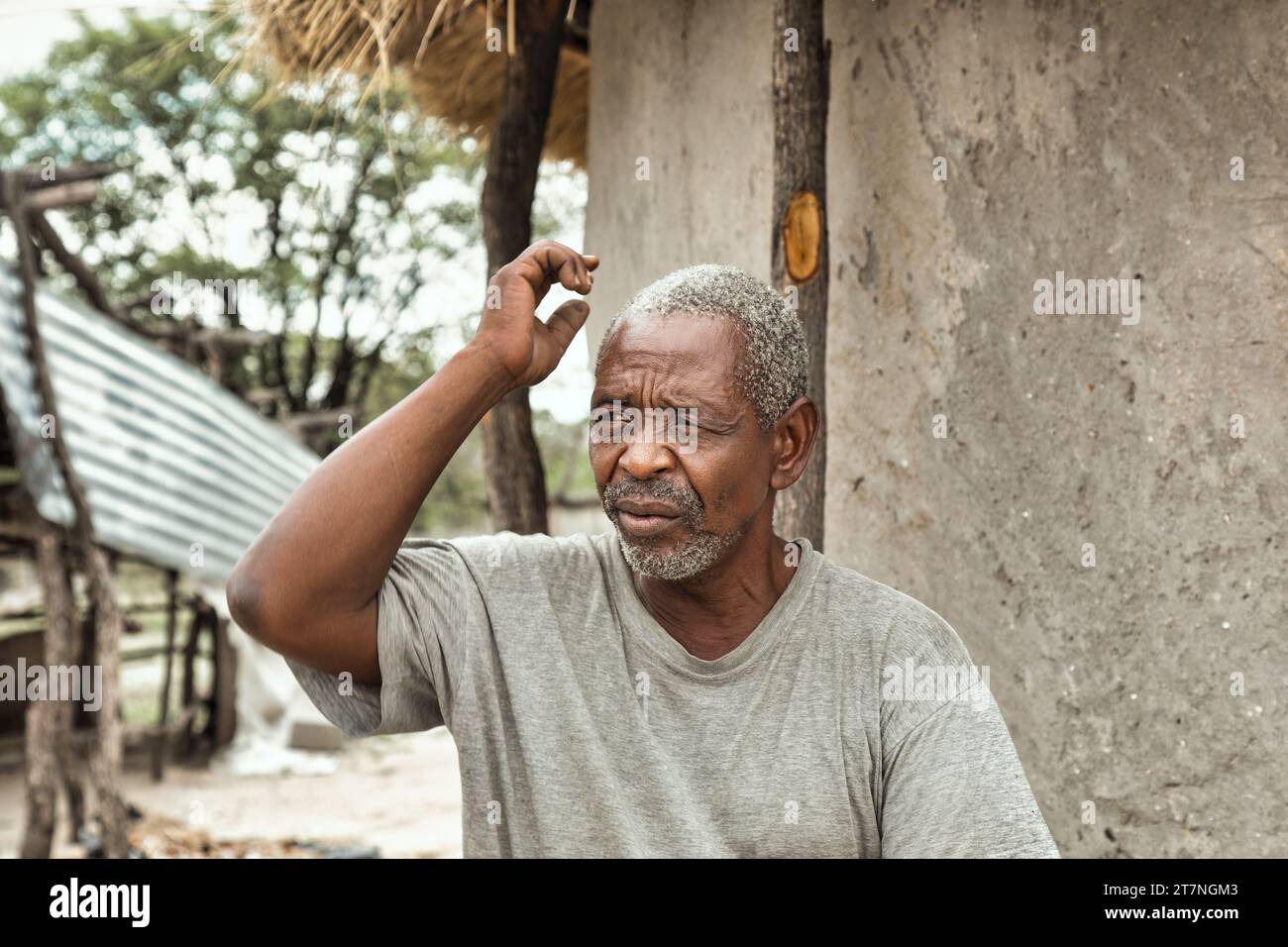 old retired african man scratching his head, some unexpected problem, in front of the house with thatched roof in the back yard in a village in africa Stock Photo