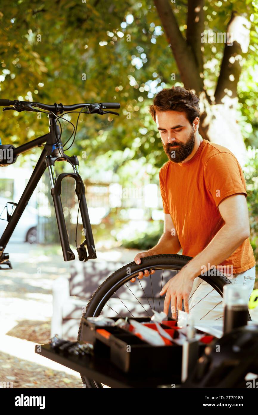 In home yard, young energetic man is using expert tools to replace damaged bike wheel. Servicing on disassembled bicycle outside for pleasure cycling is a committed, healthy caucasian male cyclist. Stock Photo