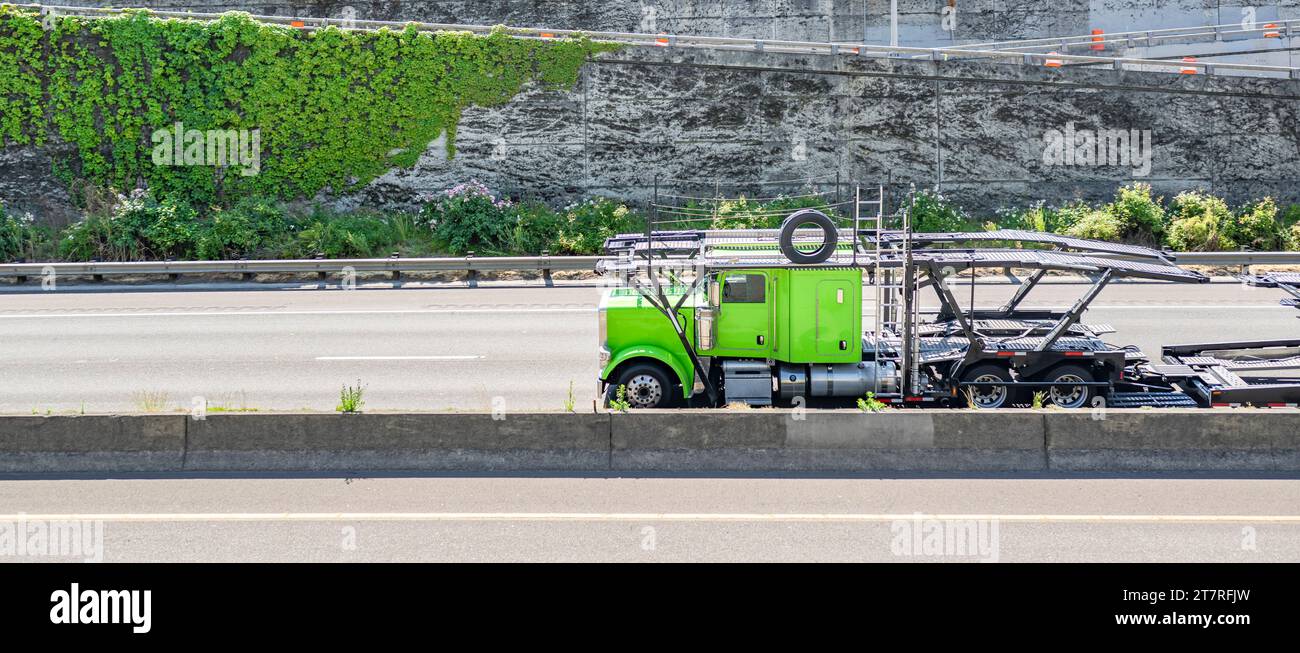 Industrial green car hauler carrier big rig semi truck tractor transporting empty hydraulic semi trailer driving on the divided highway road with conc Stock Photo