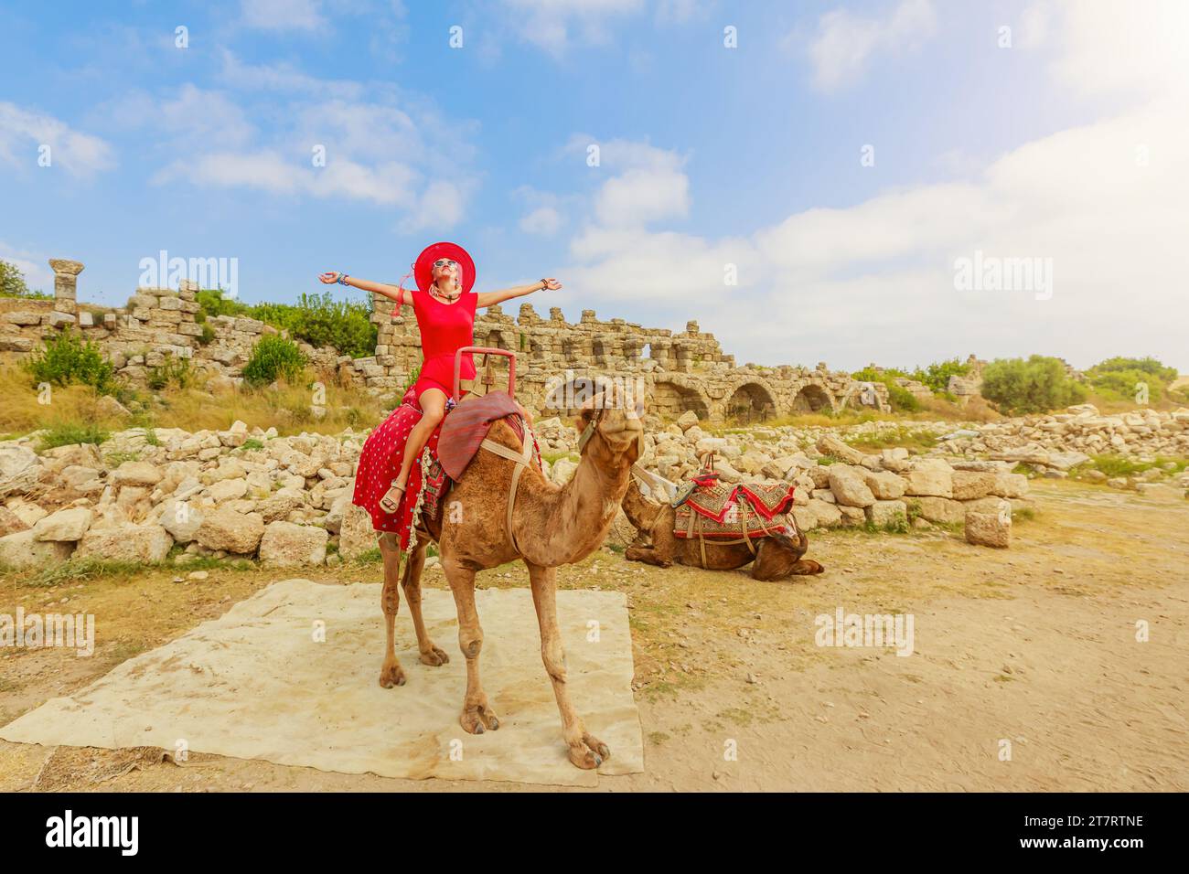 Woman sits atop a charming camel at Side archaeological site in Turkey, offering a unique perspective. Architectural remnants vividly display a fusion Stock Photo