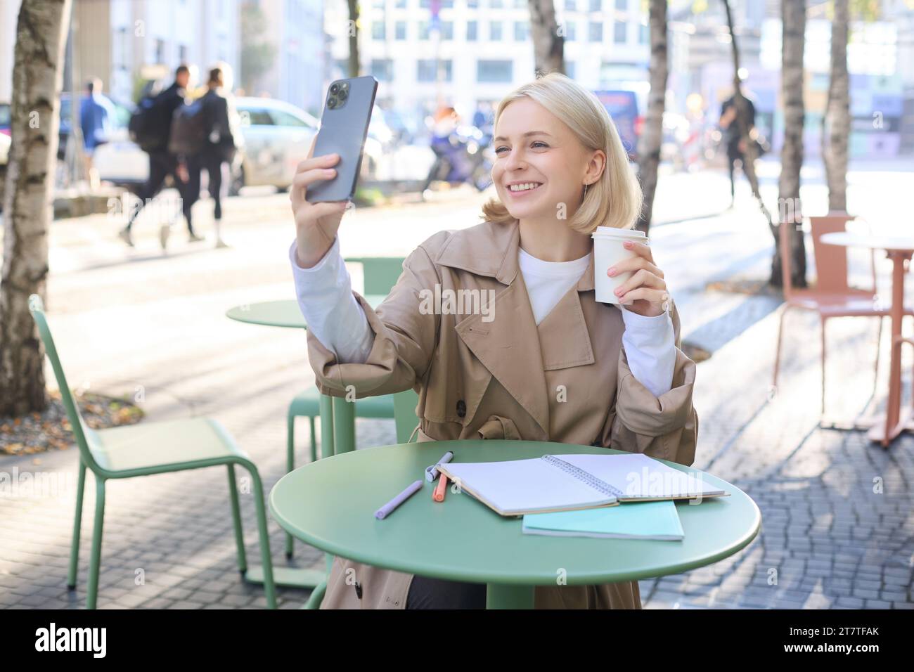 Happy blond woman taking selfie on smartphone, sitting outdoors in cafe and drinking coffee, making a post on social media to promote favourite shop Stock Photo