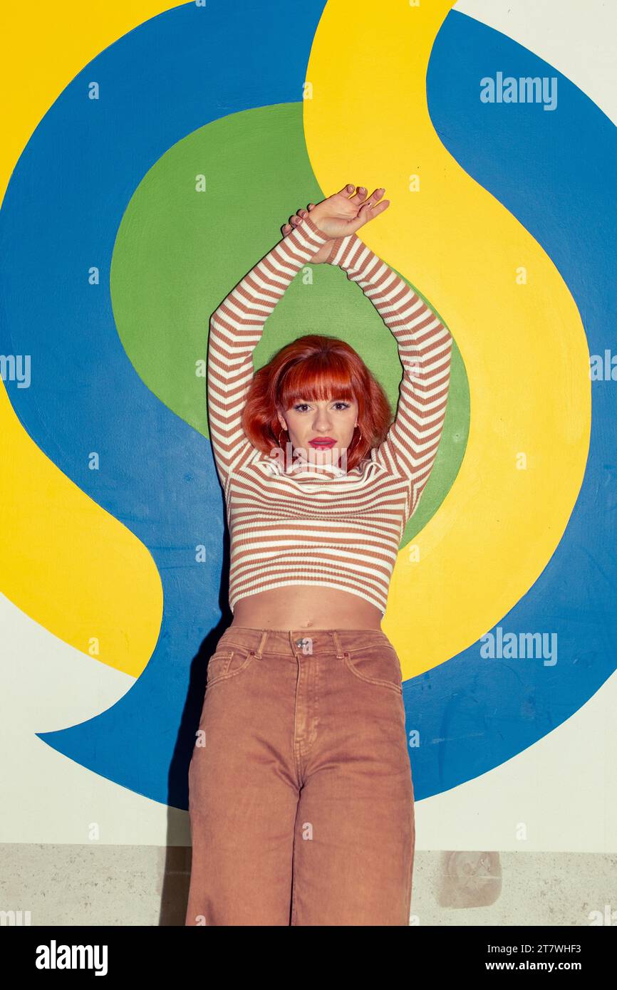Young redhead Caucasian woman in retro style of 70s posing at colorful wall both hands up Stock Photo