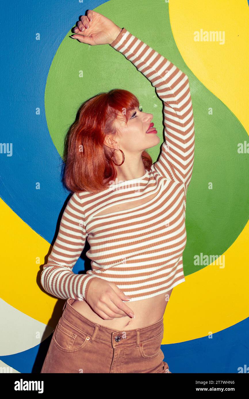 Young redhead Caucasian woman in retro style of 70s posing at colorful wall dancing vertical Stock Photo