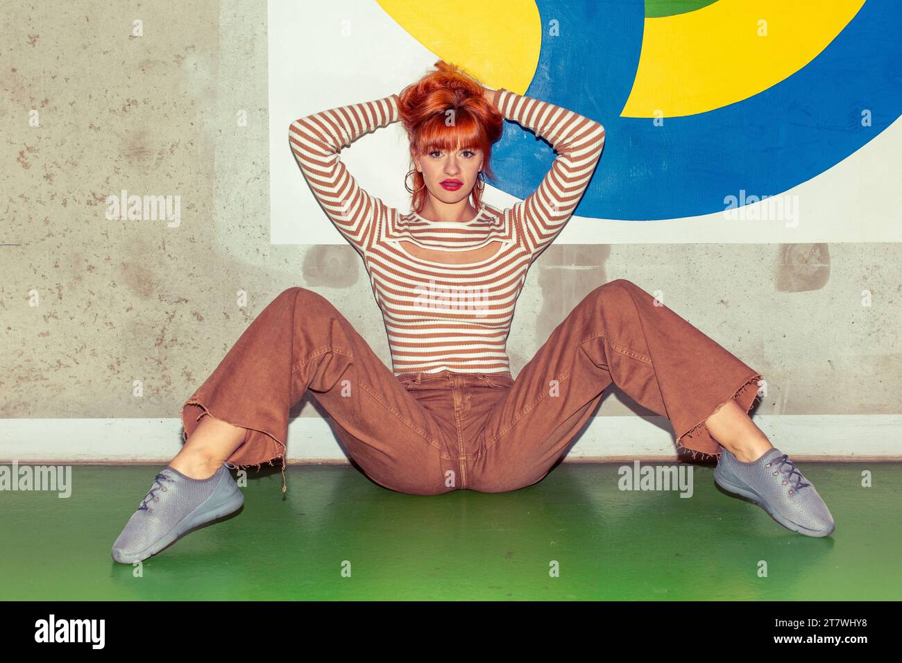 Young redhead Caucasian woman in retro style of 70s sitting at colorful wall horizontal looking into camera hands up Stock Photo
