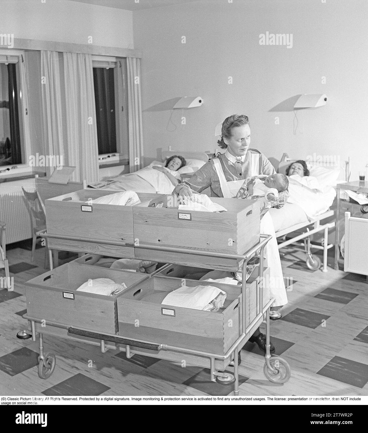 Interior from the ward for new mothers at Södersjukhuset in Stockholm where a midwife holds a newborn baby. The newborn babies lie in wooden boxes on a cart. The boxes are labeled with numbers and names. On the box that the midwife will put the baby in, it says 43 Lindberg, which is a girl who weighed 3650 grams. The new mothers lie in beds in the room. Sweden 1949. Kristoffersson ref AU33-1 Stock Photo