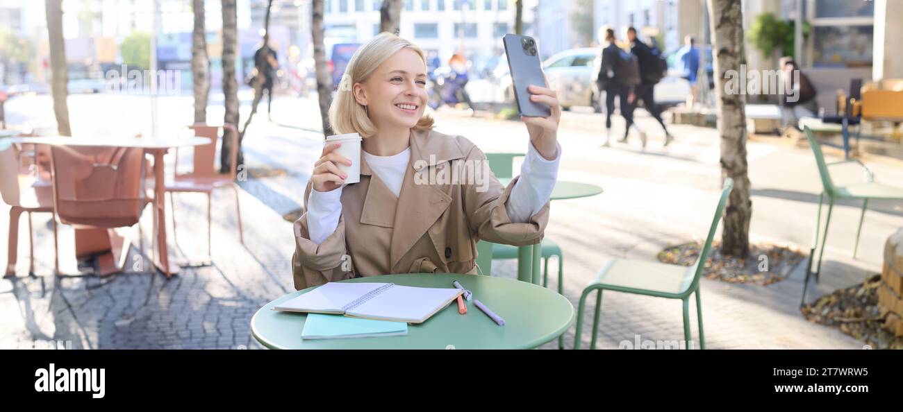 Happy blond woman taking selfie on smartphone, sitting outdoors in cafe and drinking coffee, making a post on social media to promote favourite shop Stock Photo