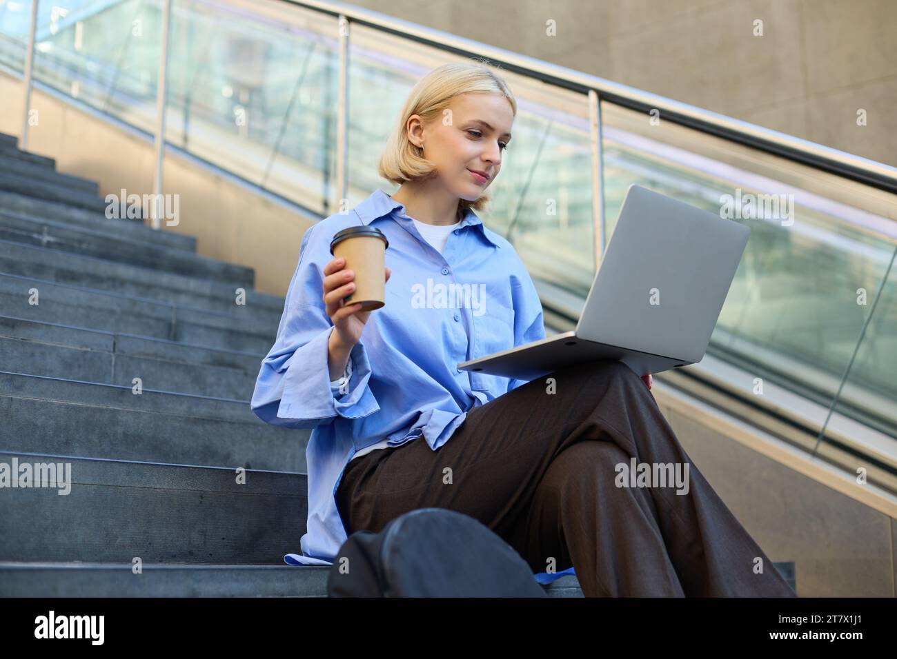Image of stylish young modern woman, student doing homework, studying outdoors on campus stairs, sitting with laptop and coffee, drinking her cappucci Stock Photo