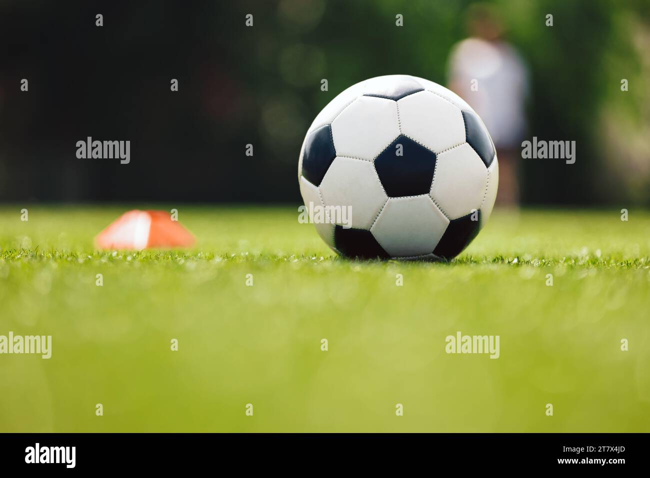 Soccer Ball and Red Cone Marker on Training Pitch. Football Turf. Soccer Stadium in Blurred Background. Young Players Practicing in the Background Stock Photo
