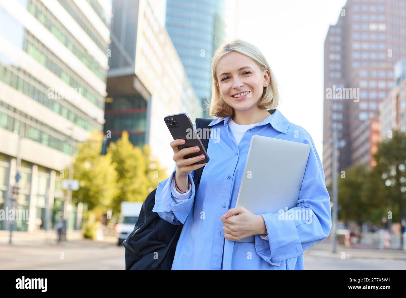 Cheerful smiling blond woman with mobile phone, posing with laptop in city centre, concept of new horizons and education opportunities. Lifestyle port Stock Photo