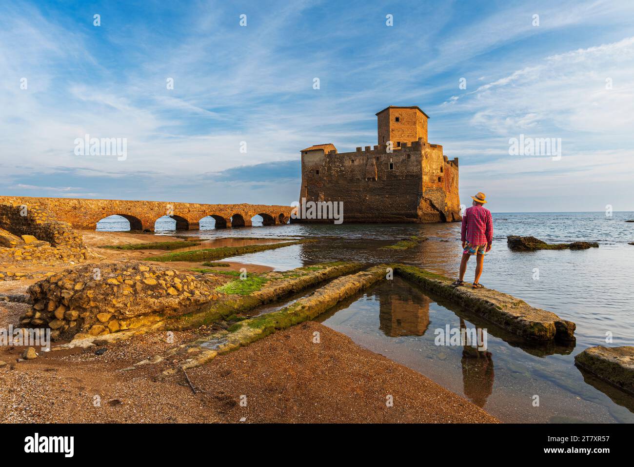 Man with hat stands on top of Roman ruin facing the medieval castle of Torre Astura rising from the sea, Nettuno municipality Stock Photo