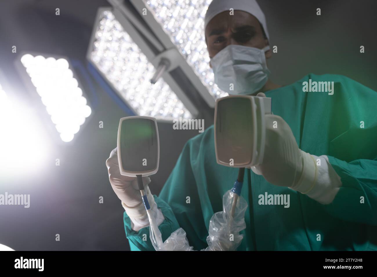 Biracial male surgeon wearing surgical gown using defibrillator in operating theatre at hospital Stock Photo