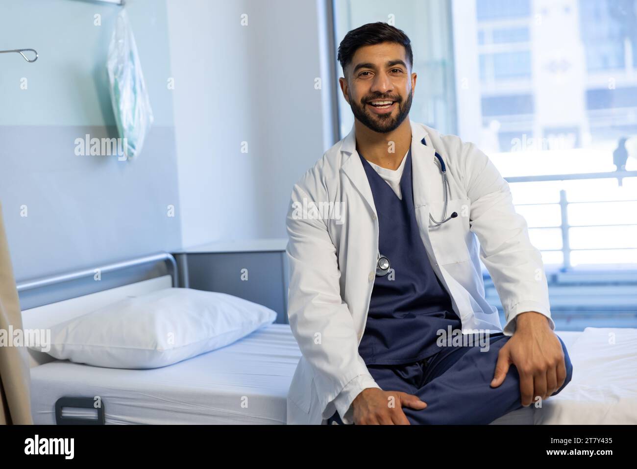 Portrait of happy biracial male doctor sitting on bed in sunny hospital ...