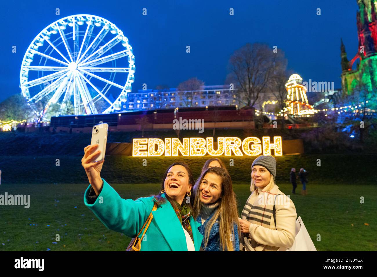 Edinburgh, Scotland, UK. 17th November, 2023. The traditional Christmas Market in East Prices Street garden opened this evening and was quickly thronged with tourists and locals. Pic; Female tourists from Newcastle pose for a selfie in front of a large illuminated sign. Iain Masterton/Alamy Live News Stock Photo