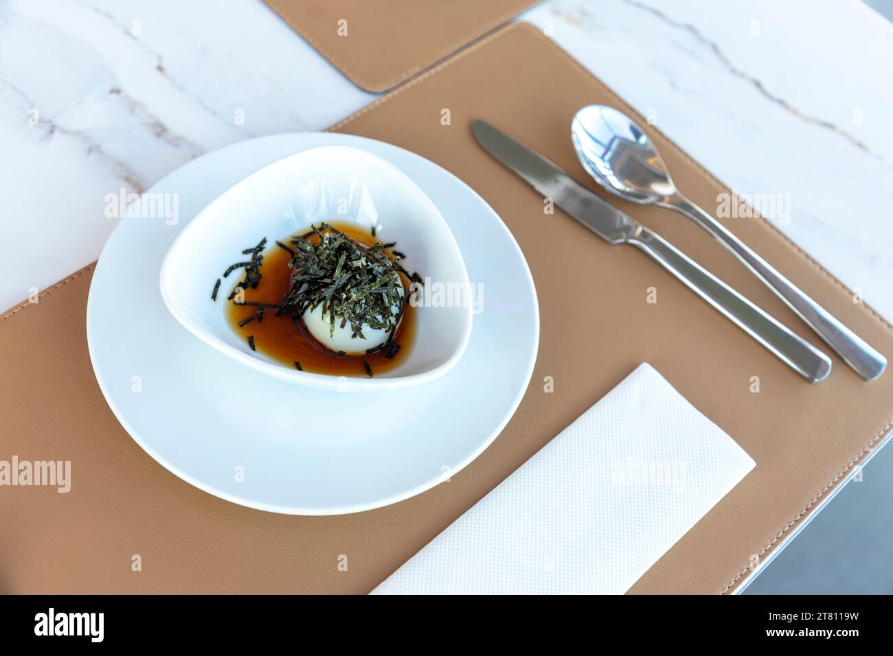 Onsen eggs soaked in sauce in a white bowl Sprinkle the top with shredded seaweed on the hotel's marble table. There is a placemat made from brown lea Stock Photo