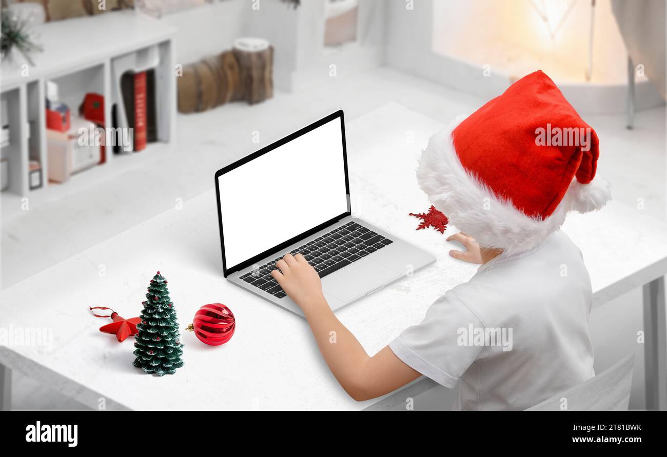 Child using laptop mockup with Christmas hat. Seasonal room with festive decorations on desk. Holiday technology concept Stock Photo
