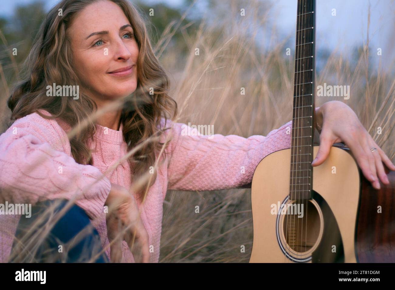 Portrait of beautiful woman musician with acoustic guitar sitting on dry grass in field. Relaxing outdoors on warm autumn evening at sunset. Attractiv Stock Photo