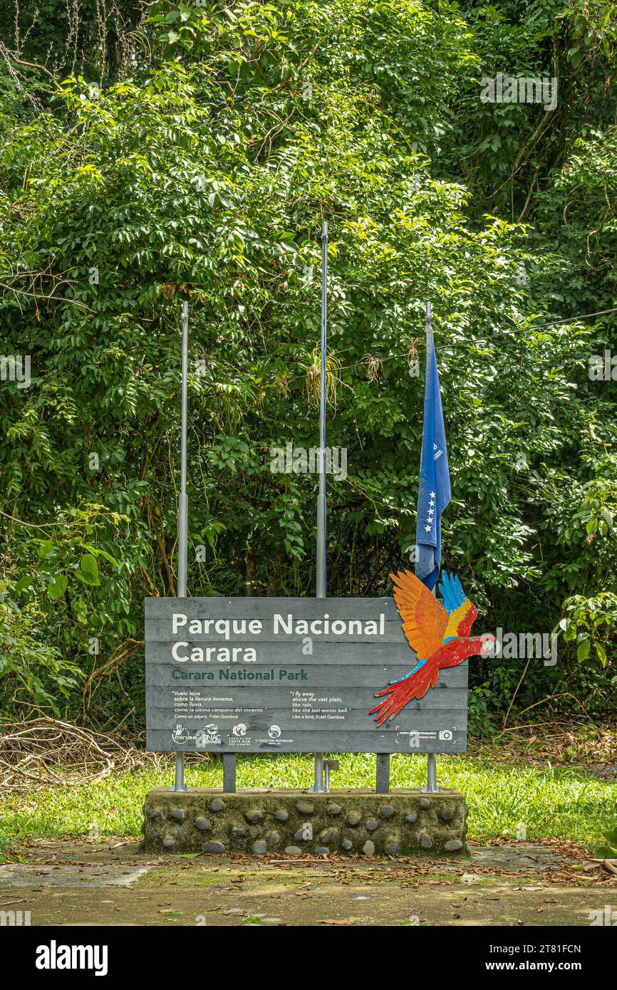 Costa Rica, Parque Nacional Carara - July 22, 2023: Closeup, official white on gray wood welcome sign with image of scarlet macaw and blue flag. Green Stock Photo