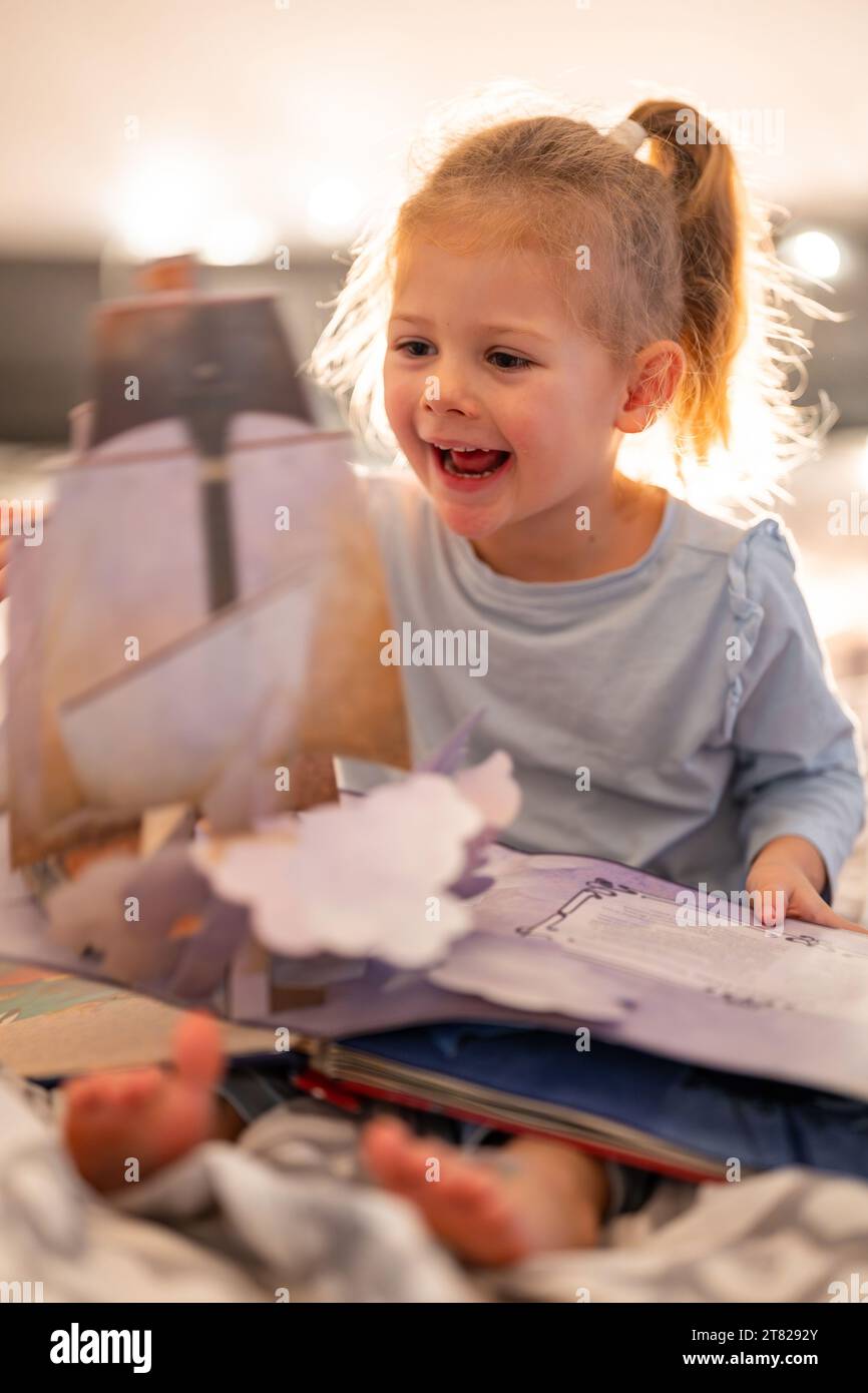 Little girl reading pop up book in home bed in Christmas environment with lights background Stock Photo