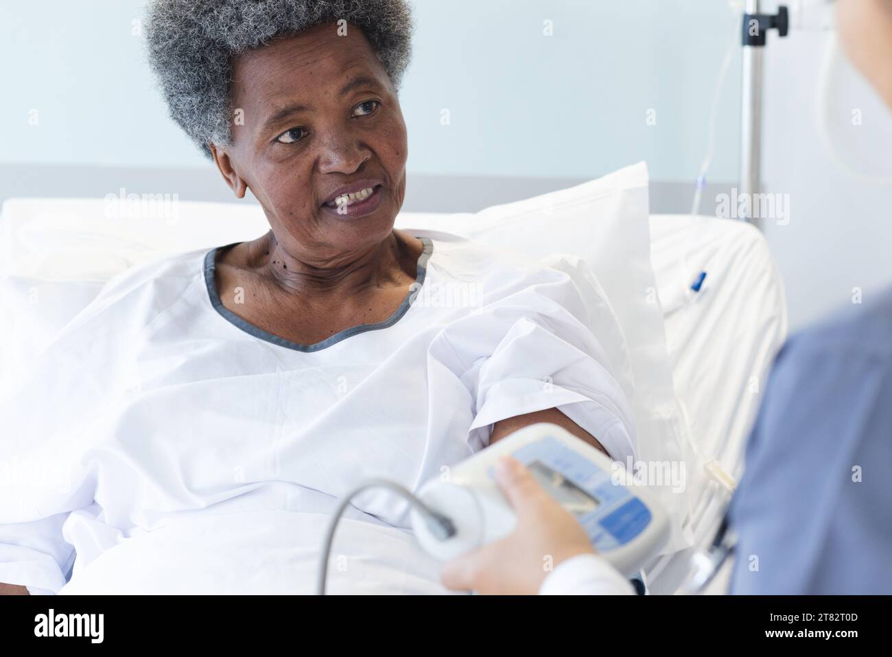 Diverse female doctor testing blood pressure of senior female patient in hospital room Stock Photo