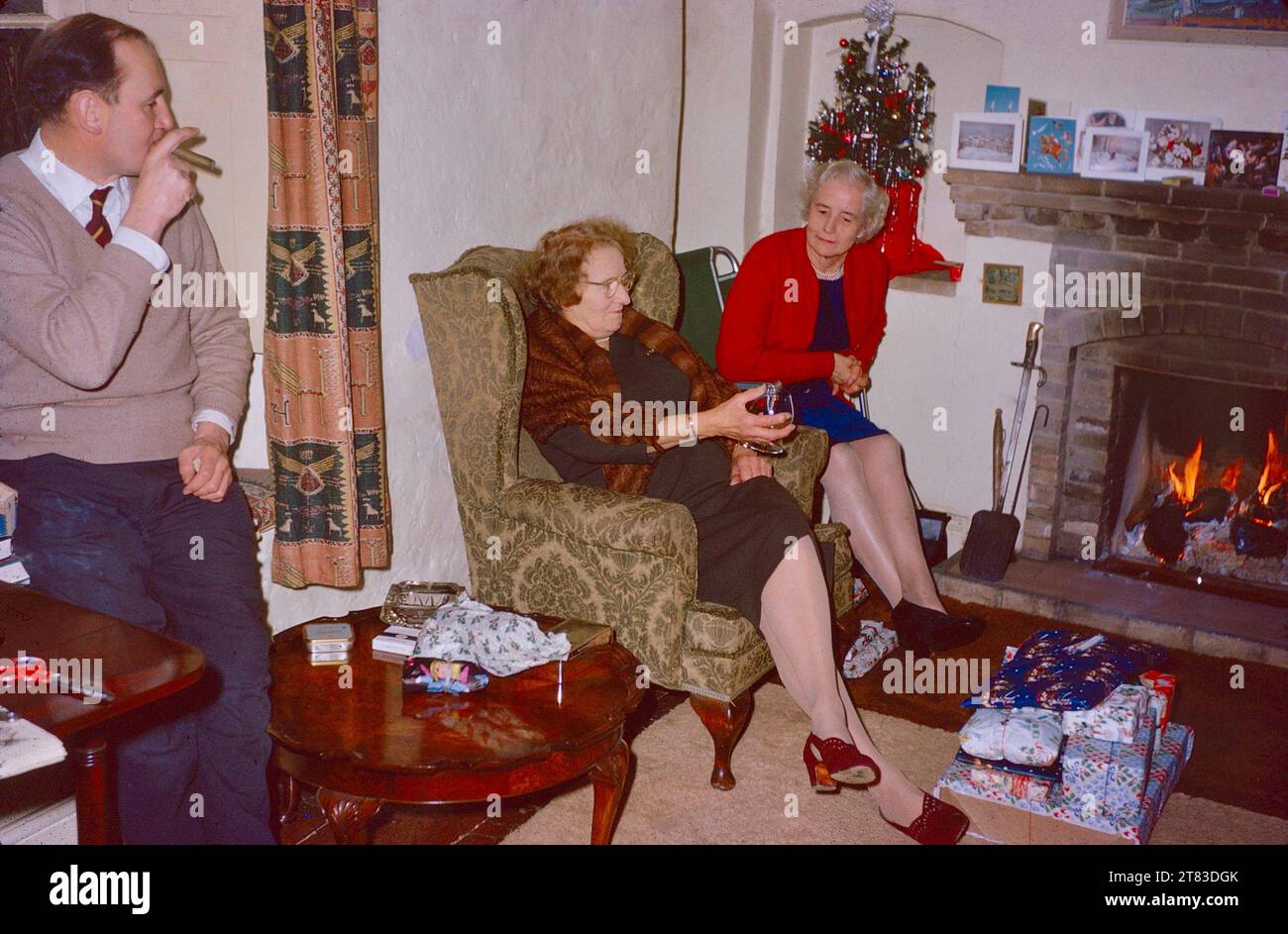 An English middle-class family group relaxing, drinking and smoking, before opening Christmas presents in 1962. Two elderly women sitting by a real log fire, one with an aperitif. A man smoking a cigar, leaning on. a window sill. A pile of Christmas presents on the floor. Christmas tree and Christmas cards in the background. Christmas before children 1960s Stock Photo