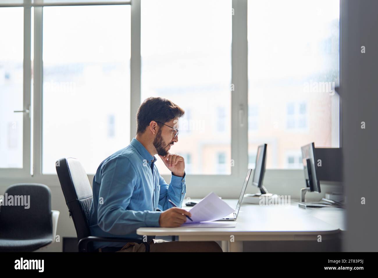 Busy serious young business man working on laptop computer holding documents. Stock Photo